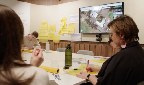 The Adelaide Design Excellence working group are sitting around a work table with yellow trace sheets pinned up behind working together on a project review. The project is on a screen above on the wall and team members are joining the meeting via a zoom call.