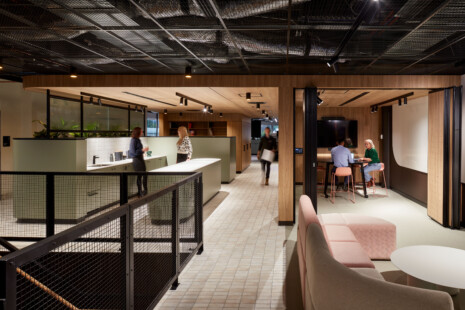 Level one of LMS Energy Headquarters arrives at a communal space with modular lounges in sand and pale pink. Sandy coloured tiles lead through a breakout kitchen in pale green, matt black fittings and fixtures, and white benches. A couple of staff gather in the kitchen chatting. Another staff member walks through from the offices at the rear with a box. Two staff meet in a meeting room off to the side that has concertina doors that open to the lounge for larger groups to gather. The warm timber joinery softens the black steel framing that separates the exposed services in the ceiling space.